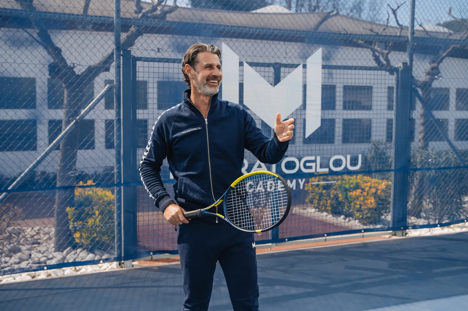 Patrick Mouratoglou smiling with a racquet in hand on an Academy court