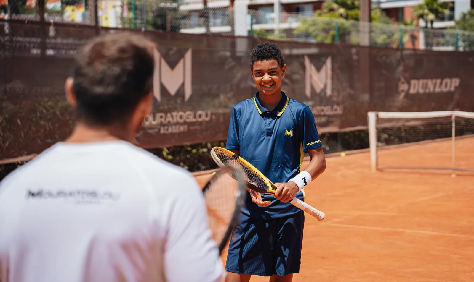 Jugador joven sonriente con su raqueta, frente a su entrenador en una pista de tierra batida