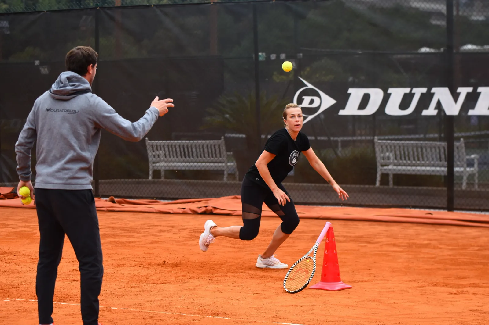 Joueuse se déplaçant vers une balle lancée par son coach sur terre battue, à côté d’un plot