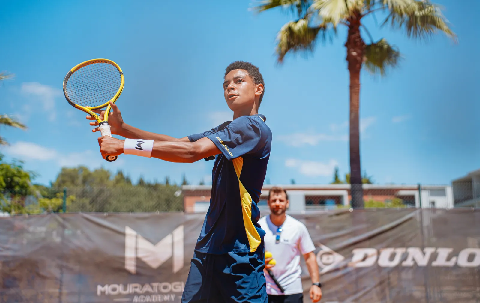 Young player finishing a two-handed backhand with a coach in the background