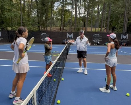 Drei Kinder am Netz eines Tennisplatzes hören den Ratschlägen ihres Coaches an der Mouratoglou Academy Atlanta zu.