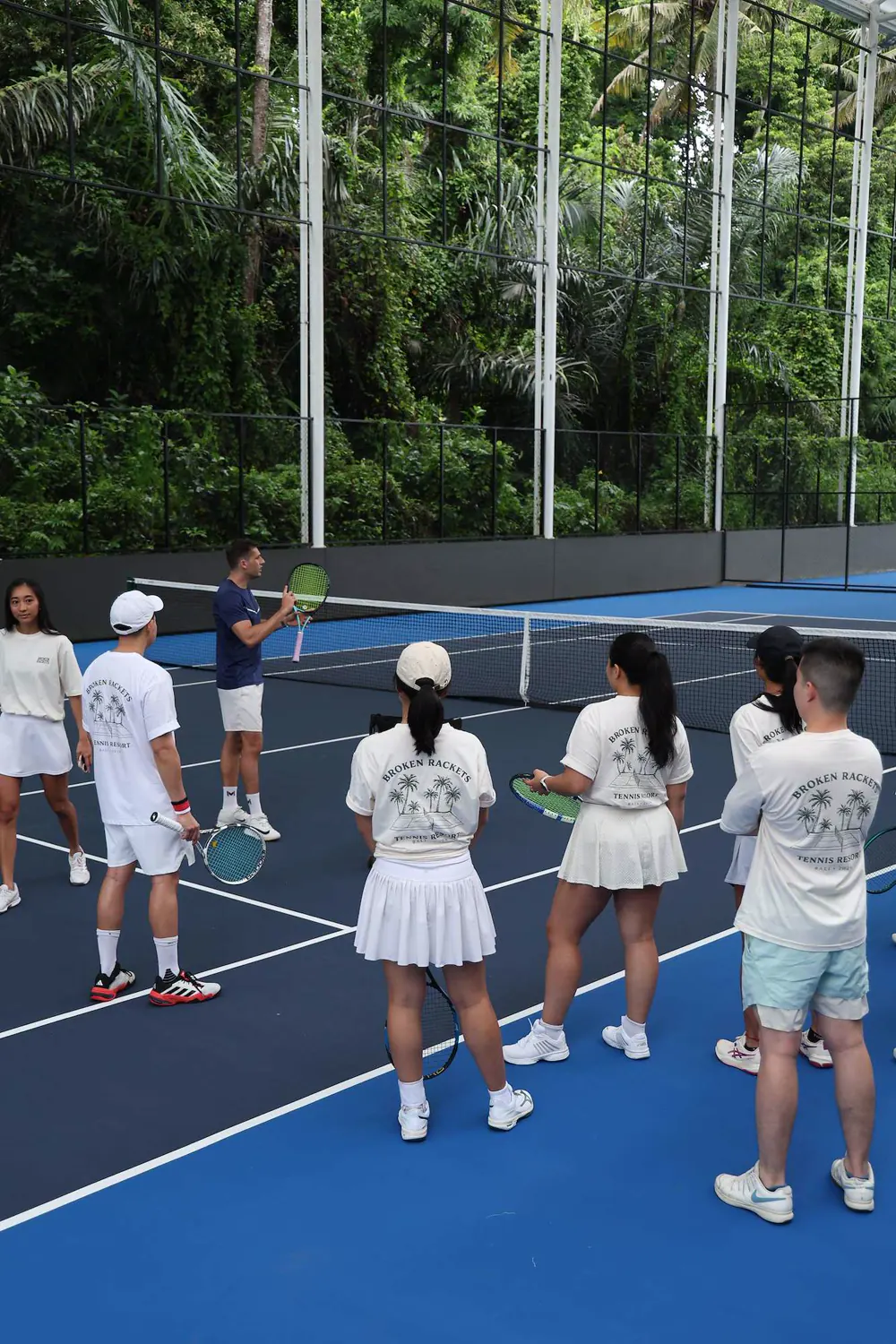 Gruppo di giocatori con magliette coordinate che ascoltano il coach durante un camp di tennis al Mouratoglou Tennis Center Bali.