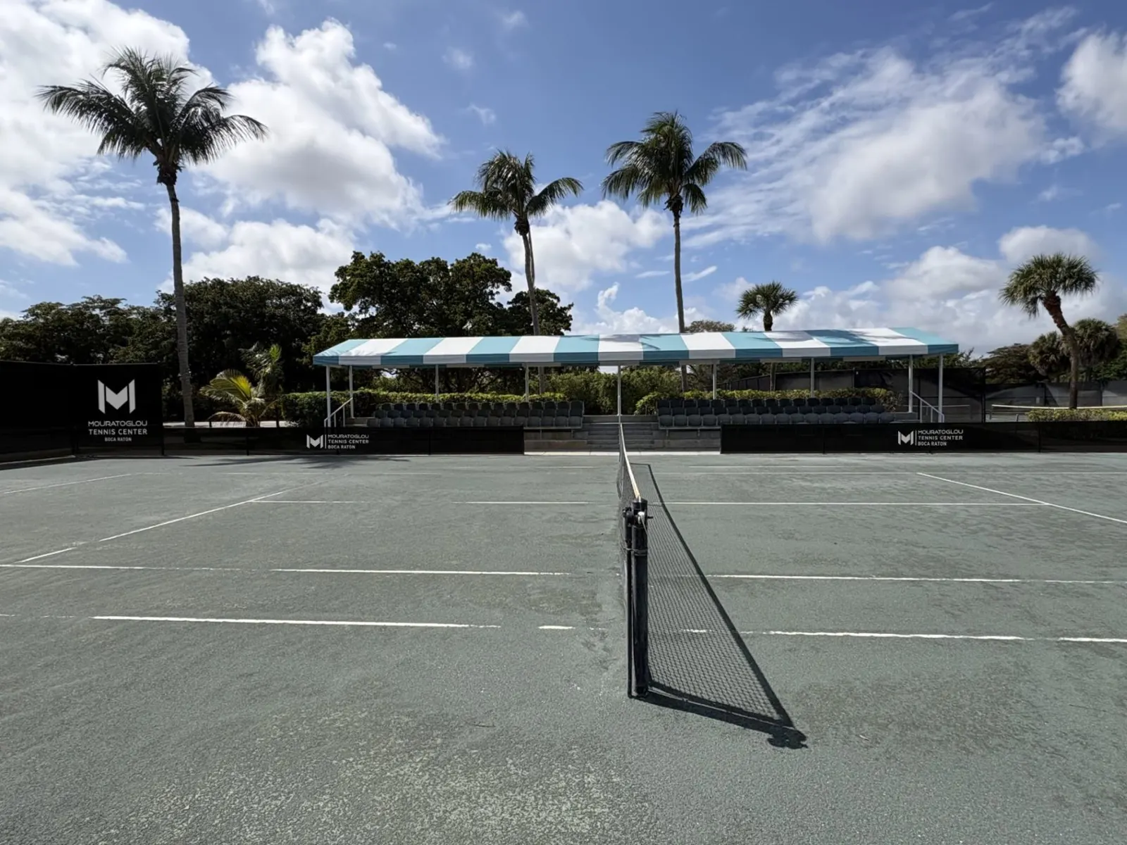 Green clay tennis court and net with palm trees and shaded seating in the background