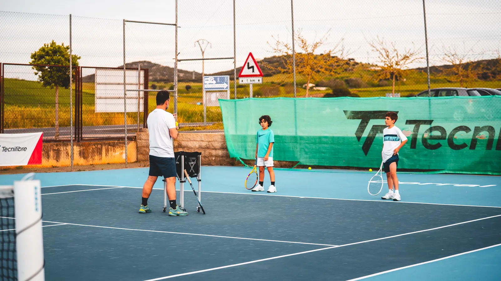 Coach mit Ballwagen trainiert zwei junge Spieler im Mouratoglou Tennis Center Calatrava.