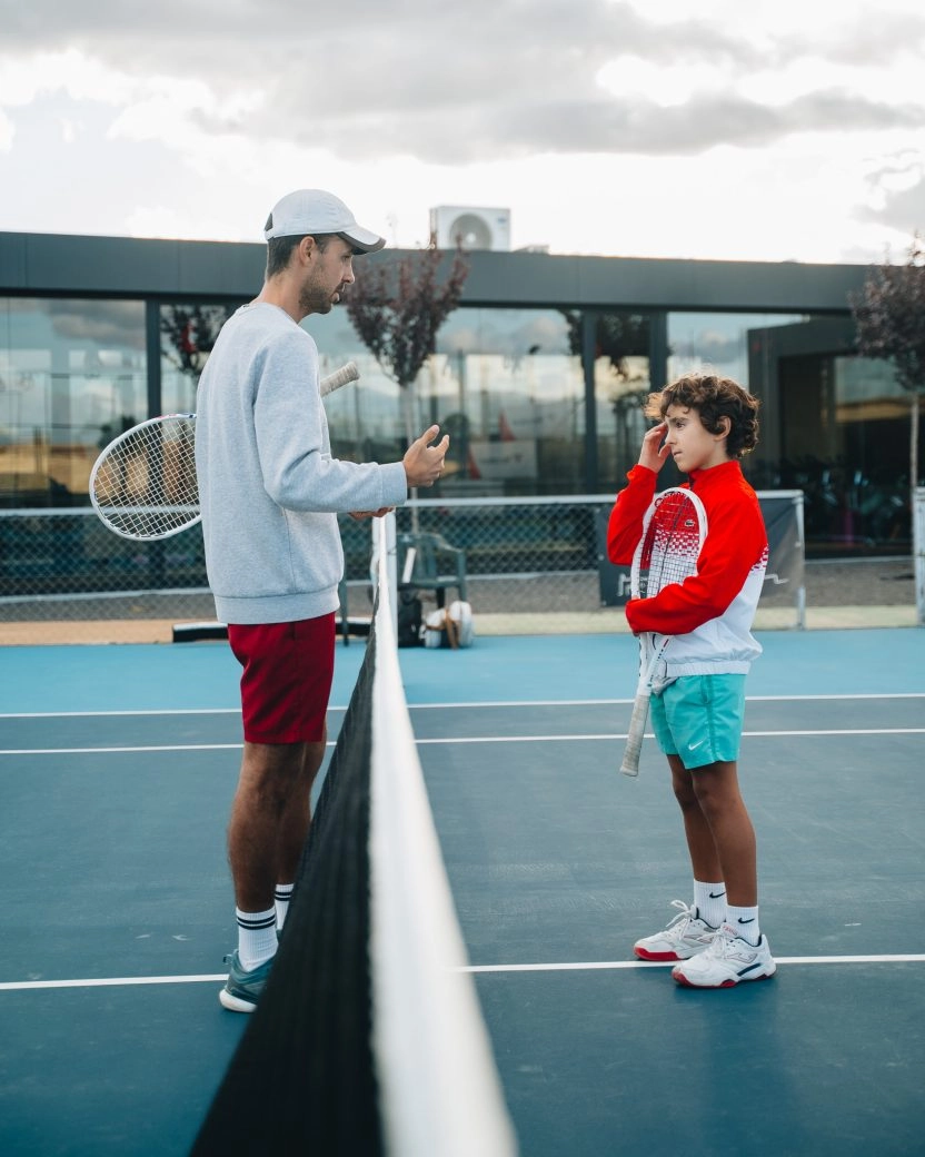 Coach im Gespräch mit einem jungen Spieler am Netz auf einem Hartplatz im Mouratoglou Tennis Center Calatrava.