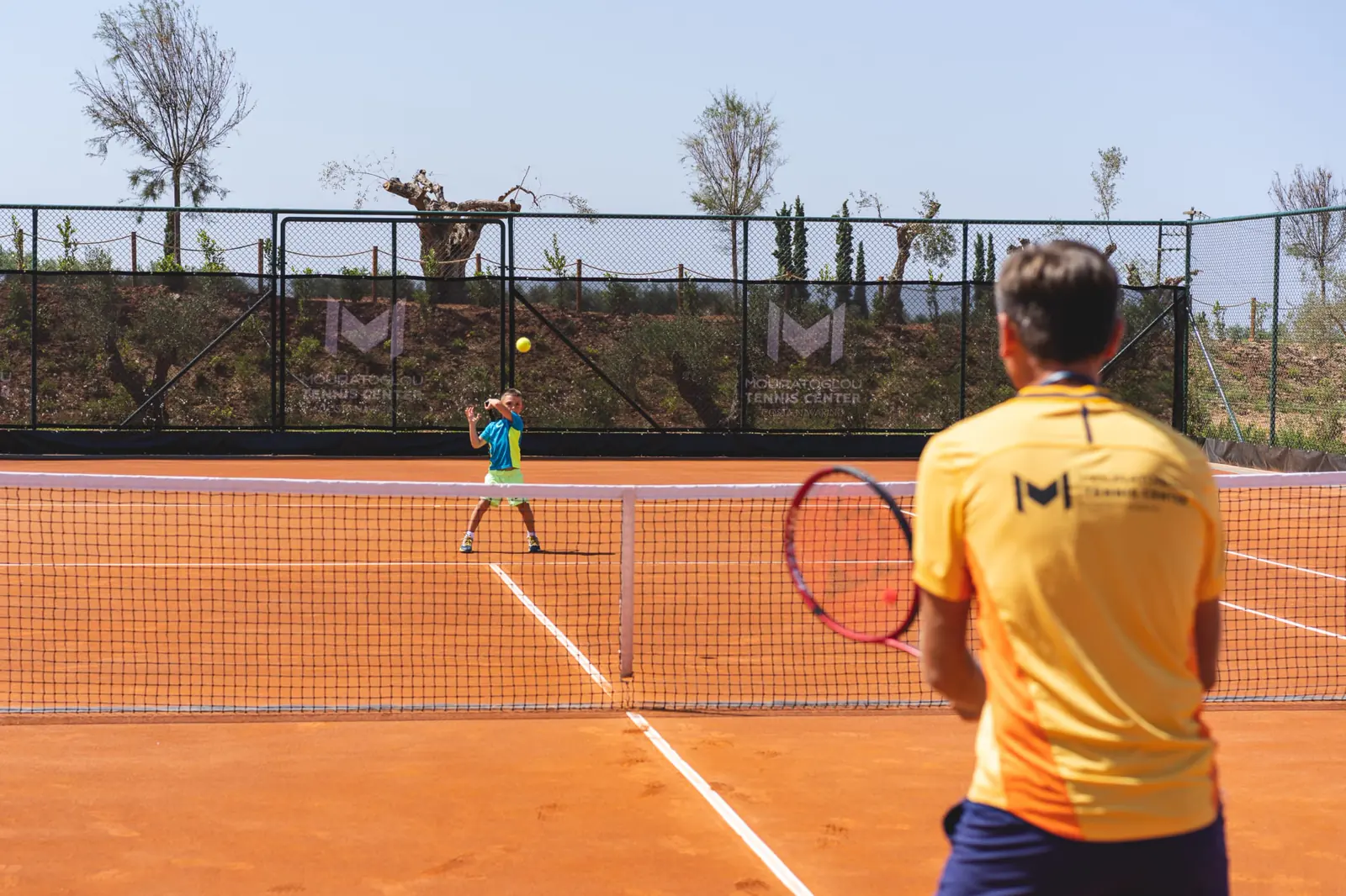 Coach Mouratoglou che allena un giovane giocatore su un campo in terra battuta durante un camp junior a Costa Navarino.