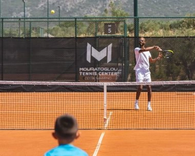 Coach che lancia palline a un giovane giocatore oltre la rete su un campo in terra battuta a Costa Navarino.