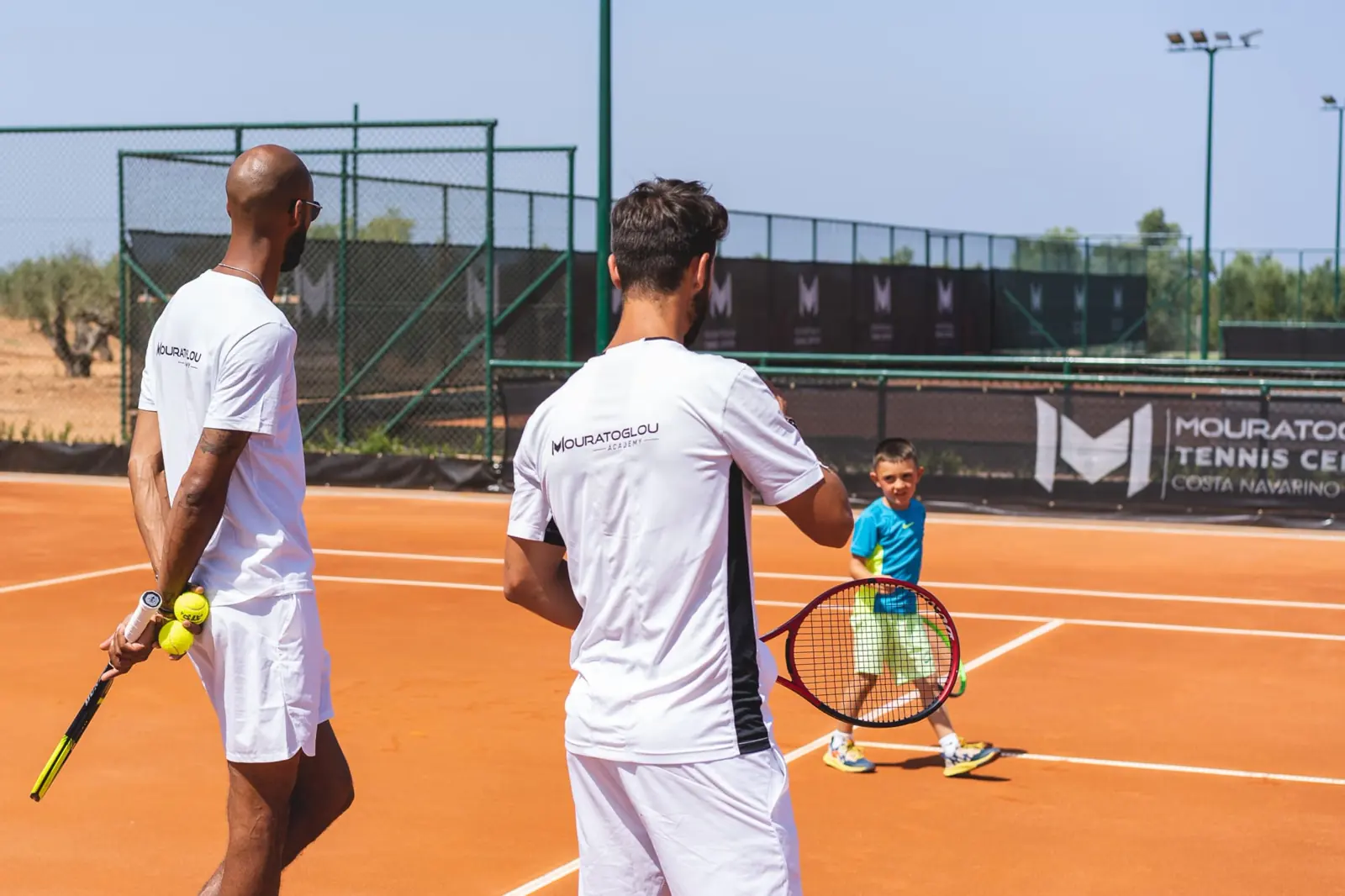 Due coach Mouratoglou con un giovane giocatore su un campo in terra battuta a Costa Navarino.