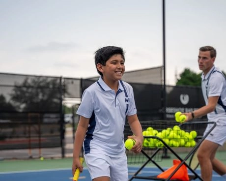 Un joven tenista sonriendo mientras entrena bajo la supervisión de un entrenador en una pista exterior del Mouratoglou Tennis Center Horseshoe Bay.