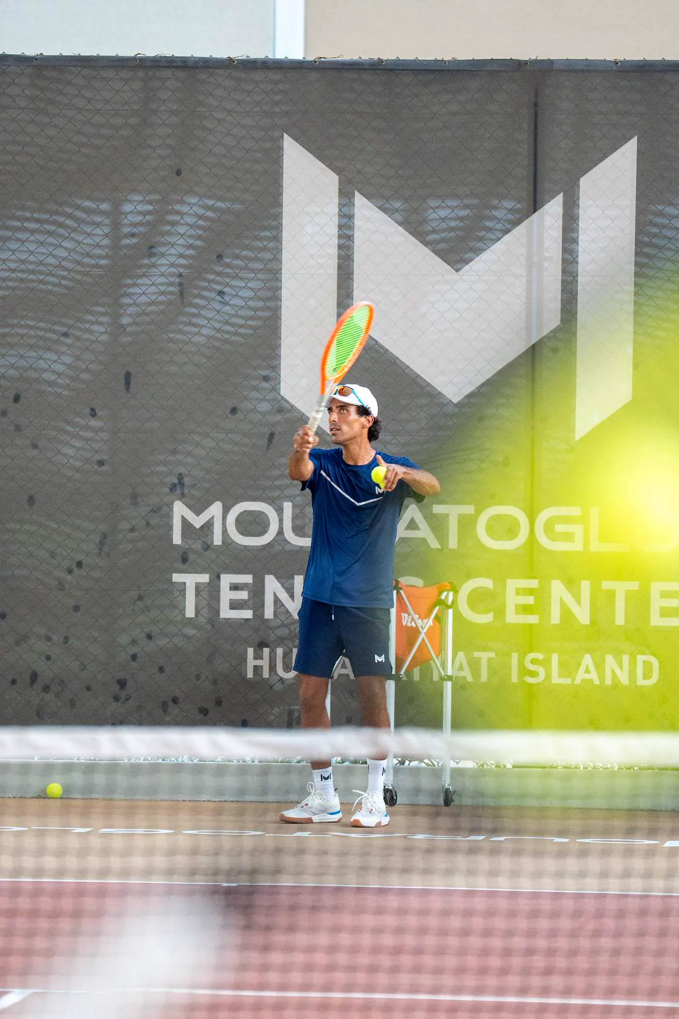 Entrenador Mouratoglou sosteniendo una pelota frente al logotipo de Mouratoglou en Abu Dabi.