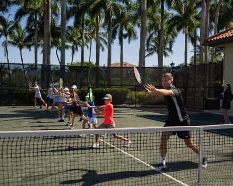 Clase de tenis en grupo para niños practicando ejercicios de derecha en una pista exterior de tierra batida con palmeras