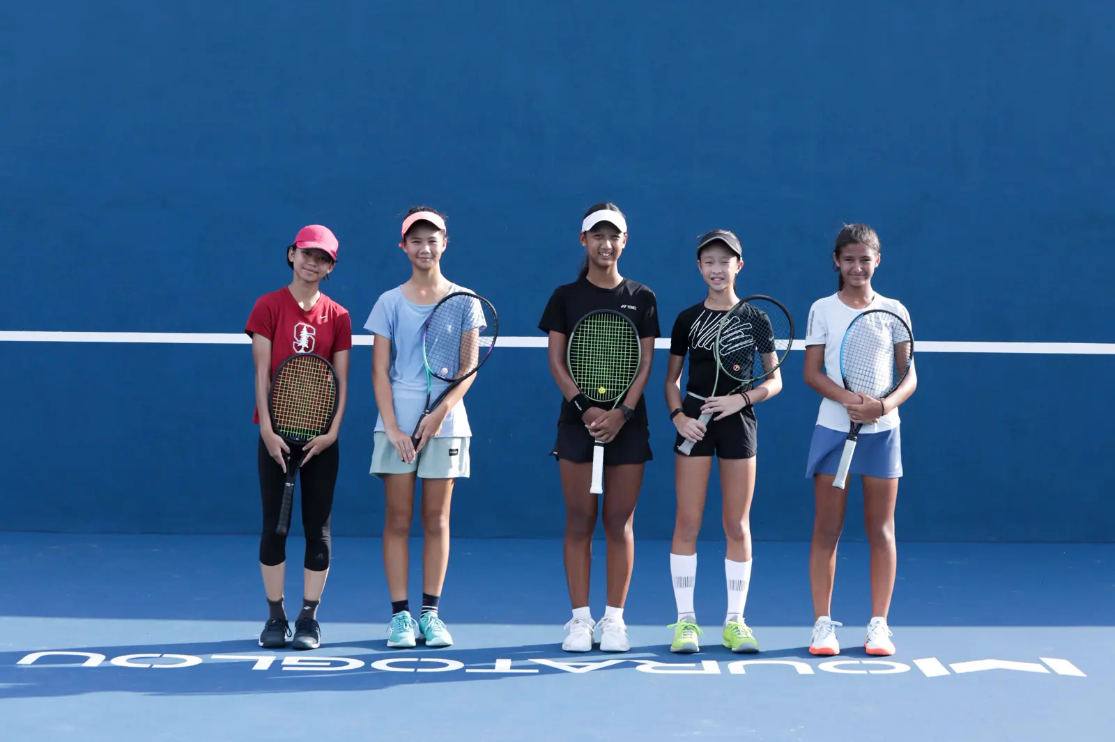 Grupo de jugadores junior de tenis de pie con sus raquetas en una pista azul de la Mouratoglou Academy Malaysia