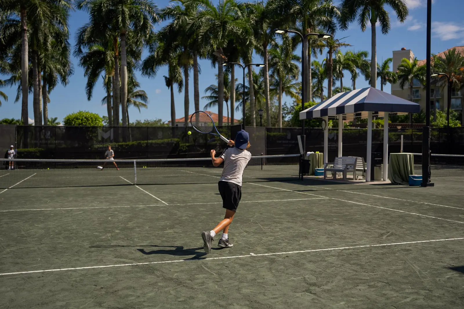 Junger Spieler schlägt eine Vorhand auf einem von Palmen umgebenen Sandplatz in Naples.