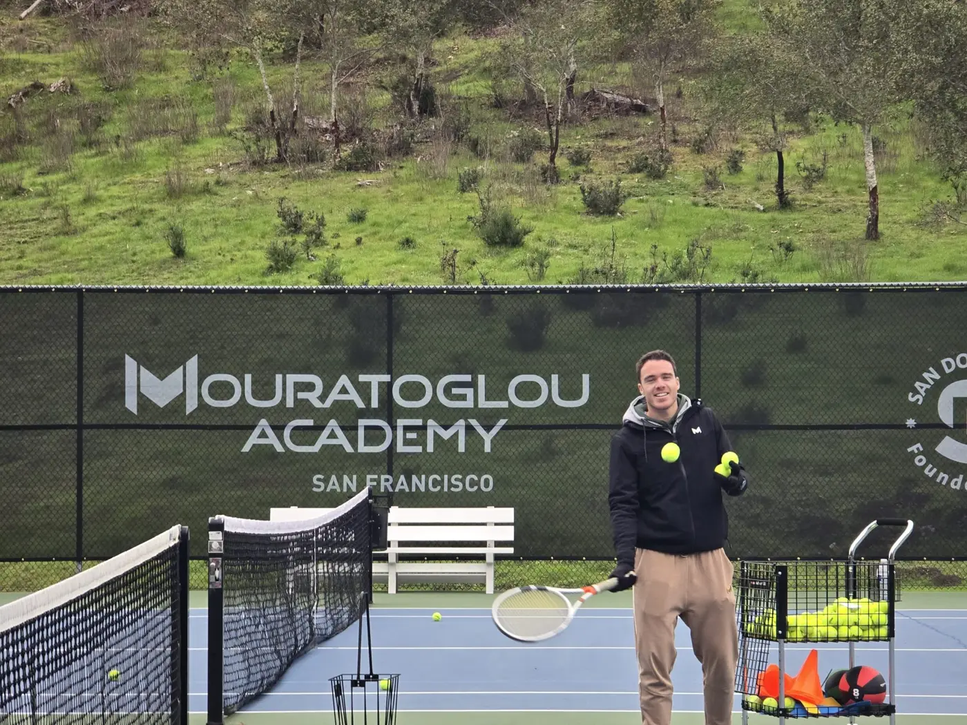 Mouratoglou coach feeding balls in front of the Mouratoglou Academy San Francisco banner