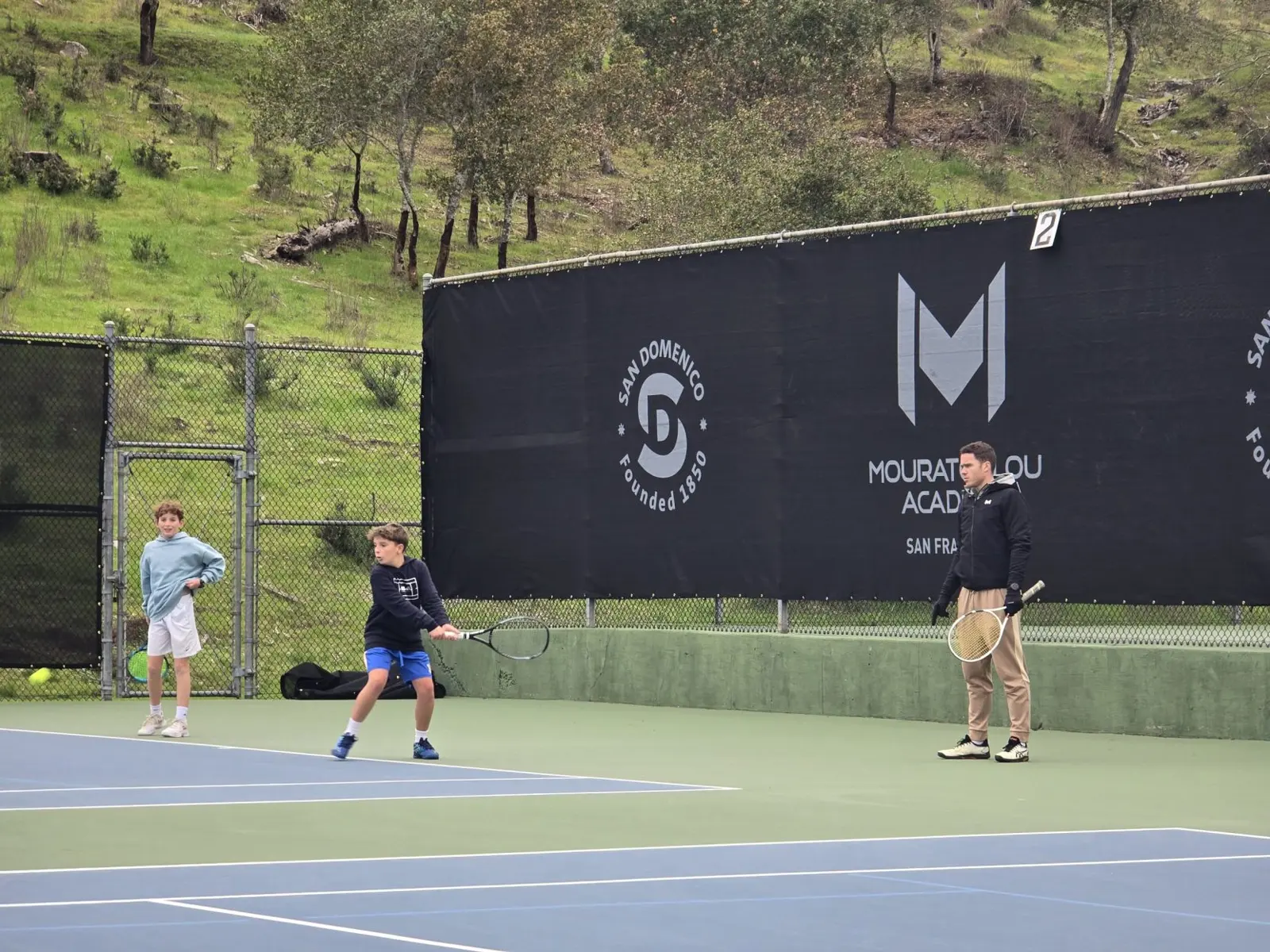 Tenniscoach trainiert zwei Jungen auf einem Außenplatz der Mouratoglou Academy San Francisco