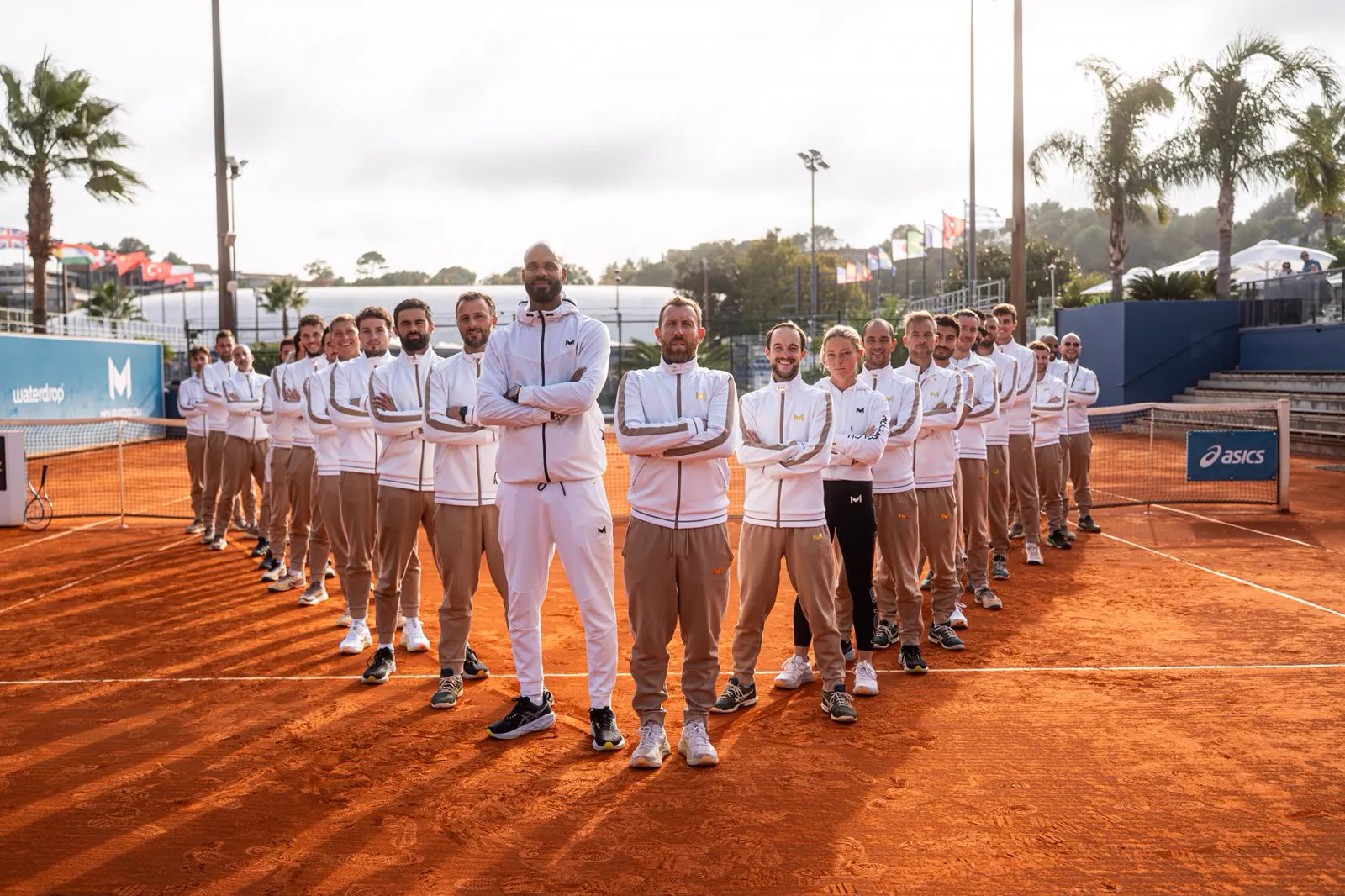 Das Trainerteam der Mouratoglou Academy in weißen und beigen Trainingsanzügen aufgereiht auf einem Sandplatz