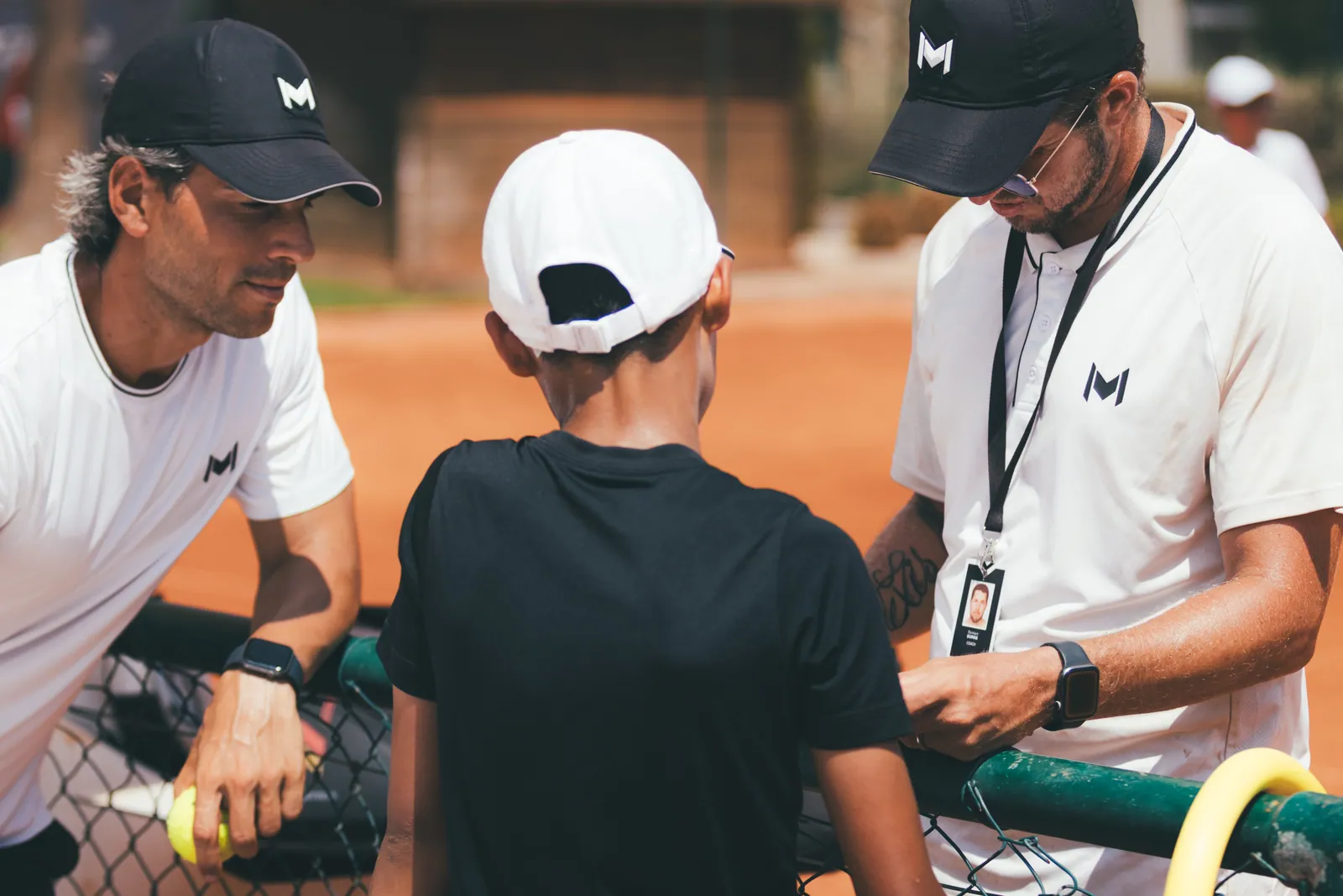 Deux coachs Mouratoglou conseillant un jeune joueur au bord d’un court en terre battue